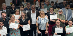 Imagem de uma reunião especial realizada na Câmara Municipal de Belo Horizonte (CMBH), em alusão ao Dia Nacional de Luta da Pessoa com Deficiência. Na foto, ao centro, está o vereador de BH Wagner Ferreira (homem negro, alto de camisa branca e paletó preto) e Sônia Souza (mulher branca, baixa, loira apoiada em muletas canadenses) representando o Núcleo da Pessoa com Deficiência (NPD) do SINJUS-MG junto a outros homenageados por sua atuação em prol da inclusão que estão posando em pé e segurando certificados de reconhecimento.