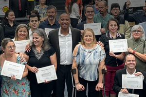 Imagem de uma reunião especial realizada na Câmara Municipal de Belo Horizonte (CMBH), em alusão ao Dia Nacional de Luta da Pessoa com Deficiência. Na foto, ao centro, está o vereador de BH Wagner Ferreira (homem negro, alto de camisa branca e paletó preto) e Sônia Souza (mulher branca, baixa, loira apoiada em muletas canadenses) representando o Núcleo da Pessoa com Deficiência (NPD) do SINJUS-MG junto a outros homenageados por sua atuação em prol da inclusão que estão posando em pé e segurando certificados de reconhecimento.