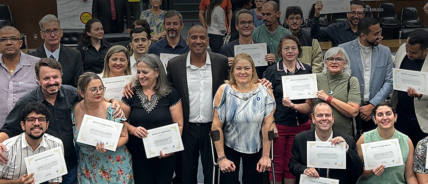 Imagem de uma reunião especial realizada na Câmara Municipal de Belo Horizonte (CMBH), em alusão ao Dia Nacional de Luta da Pessoa com Deficiência. Na foto, ao centro, está o vereador de BH Wagner Ferreira (homem negro, alto de camisa branca e paletó preto) e Sônia Souza (mulher branca, baixa, loira apoiada em muletas canadenses) representando o Núcleo da Pessoa com Deficiência (NPD) do SINJUS-MG junto a outros homenageados por sua atuação em prol da inclusão que estão posando em pé e segurando certificados de reconhecimento.